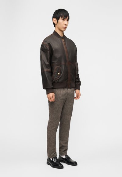 Άνδρας στέκεται φορώντας σκούρο καφέ δερμάτινο μπουφάν τύπου bomber, γκρι μάλλινες παντελόνες, λευκές κάλτσες και μαύρα δερμάτινα loafers, μπροστά σε λευκό απλό φόντο.