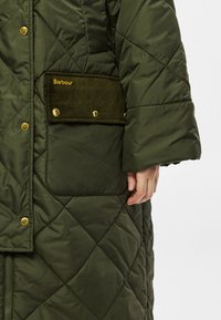 Zelený prošívaný kabát s manšestrovou kapsou se zlatě vyšitým logem, velkými knoflíky a zapínáním na zip. Texturovaná látka a detaaily designu.