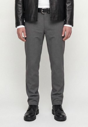 Homme portant un pantalon gris à carreaux, une ceinture en cuir noire, une veste en cuir noire et des bottes noires à lacets, debout devant un fond uni.