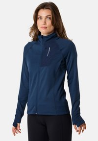 Marinblå zip-up jacka i texturerad tyg, med en horisontell ficka på bröstet och tumhål vid ärmsluten för extra komfort.