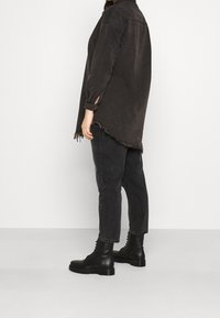 Camisa de denim negra de gran tamaño con dobladillo deshilachado y detalles en los puños, combinada con pantalones negros holgados y botas de combate negras, vista de lado.