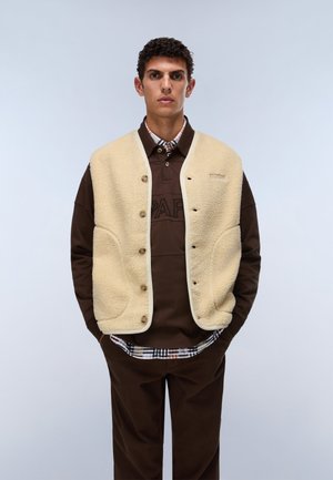 Mann trägt eine beige Fleeceweste über einem braunen Hemd mit Knöpfen und dunkelbrauner Hose, steht vor einem schlichten hellen Hintergrund.
