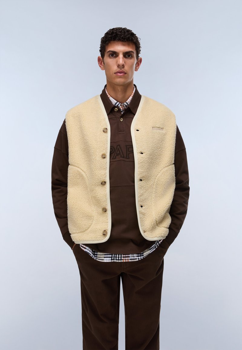 Hombre con un chaleco beige de forro polar encima de una camisa marrón con botones y pantalones marrón oscuro, de pie frente a un fondo claro y liso.