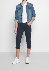 Chaqueta de mezclilla en azul claro, camisa blanca de botones, pantalones cortos navy y zapatillas blancas con suelas marrones, de pie contra un fondo neutral.