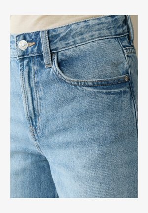 Prim-plan cu o persoană purtând blugi de denim bleu deschis, cu buzunar frontal vizibil, buclă pentru curea și nasture la talie.