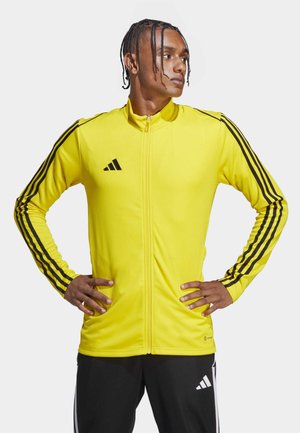 Chaqueta deportiva amarilla con un alto cuello, cremallera completa, acentos en negro con tres rayas en las mangas y un pequeño logo negro en el pecho.
