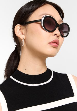Donna con capelli scuri legati indietro che indossa grandi occhiali da sole rotondi neri, orecchini a cerchio argentati e un top nero senza maniche a righe bianche.