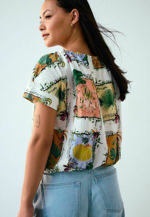 Woman with long dark hair wears short-sleeve blouse with colorful botanical prints and light blue high-waisted jeans, standing sideways.