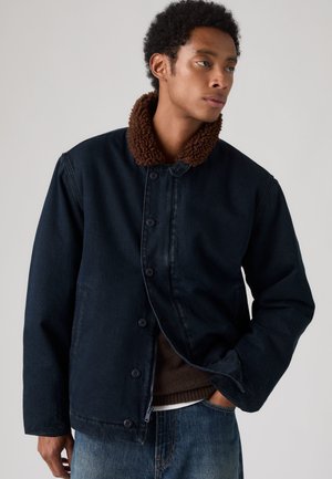 Joven con chaqueta azul oscuro con cuello de forro polar marrón, parcialmente abotonada, de pie con una mano en el bolsillo vaquero, mirando hacia la derecha.