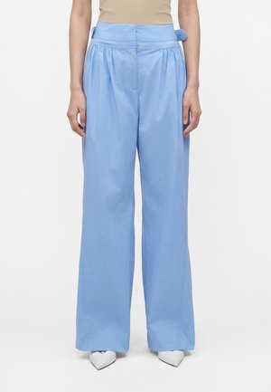 Kobieta nosząca jasnoniebieskie spodnie z wysokim stanem i szerokimi nogawkami z bocznymi detalami w pasie, zestawione z białymi, spiczastymi butami i beżowym topem.