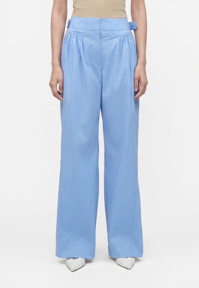 Femme portant un pantalon bleu clair taille haute à jambes larges avec des détails de ceinture sur le côté, associé à des chaussures blanches pointues et un haut beige.