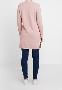 Tunika rosa a coste con maniche lunghe, fessure laterali e scollatura rotonda, abbinata a jeans skinny blu scuro e sneakers bianche.