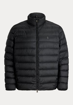 Homme portant un bonnet noir, une doudoune noire, un pantalon cargo camouflage, des bottes marron et une montre-bracelet, debout devant un fond uni.