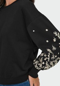 Črna sweatshirt s kontrastno cvetlično vezenino na rokavih, z zaobljenim ovratnikom in rebrastimi manšetami. Mehka bombažna tkanina.