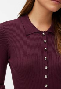 Artimos moters, dėvinčios tamsiai violetinį, briaunuotą viršutinį drabužį su apykakle ir vertikalia eile iš septynių blizgių sidabrinių sagų, vaizdas.