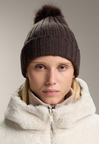 Knitted brown beanie with a fuzzy pom-pom atop, worn with a beige zip-up fleece jacket. Features ribbed texture and snug fit.