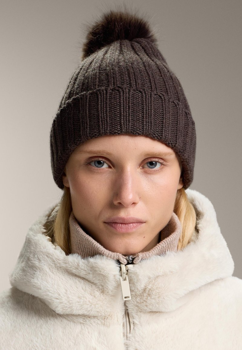 Knitted brown beanie with a fuzzy pom-pom atop, worn with a beige zip-up fleece jacket. Features ribbed texture and snug fit.
