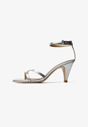 Srebrny sandał na wysokim obcasie z cienkimi paskami na palcach i wokół kostki, ozdobiony małą klamrą i zwężającym się obcasem o średniej wysokości.