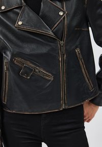 Giacca da motociclista in pelle nera con numerose tasche con zip, bordi consumati e accessori in metallo argentato. Caratterizzata da un ampio rever e chiusura frontale con zip.