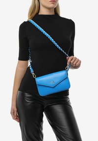 Borsa a tracolla in pelle blu con design a busta, tracolla intrecciata e dettagli in metallo argentato. Indossata sopra un outfit scuro.