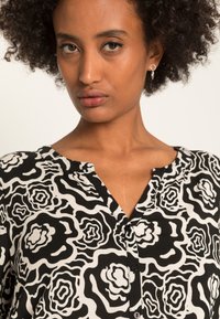 Blusa preta e branca com um padrão floral em espiral, fechamento frontal por botões e gola. Tecido leve com uma textura suave.