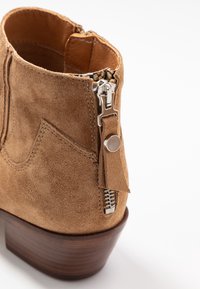 Kotníková bota z béžového semiše se zipem, koženou podšívkou a dřevěným podpatkem. Má dekorativní vzor švů vedle zipu.