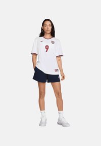 Maglia da calcio bianca con accenti rossi e numero 9, abbinata a pantaloni corti blu scuro. Indossata con calze bianche e scarpe da ginnastica, con scollatura rotonda.