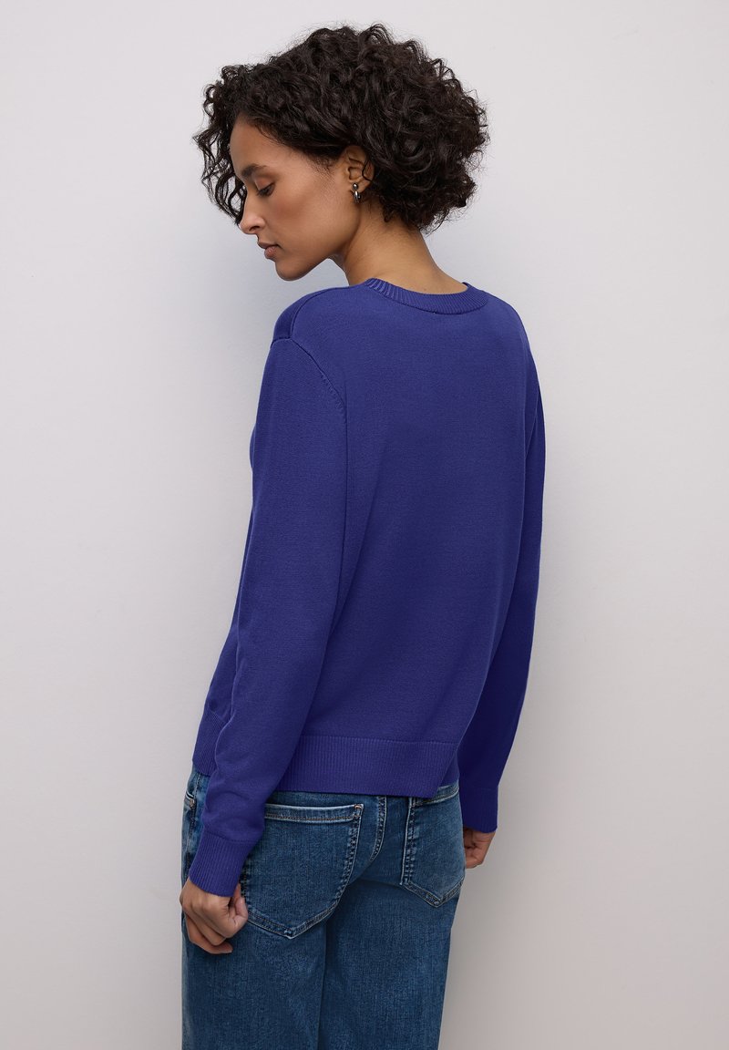 Pull en maille violet avec un col rond et des poignets côtelés, associé à un jean à jambes larges bleu clair, vu de dos sur un fond neutre.