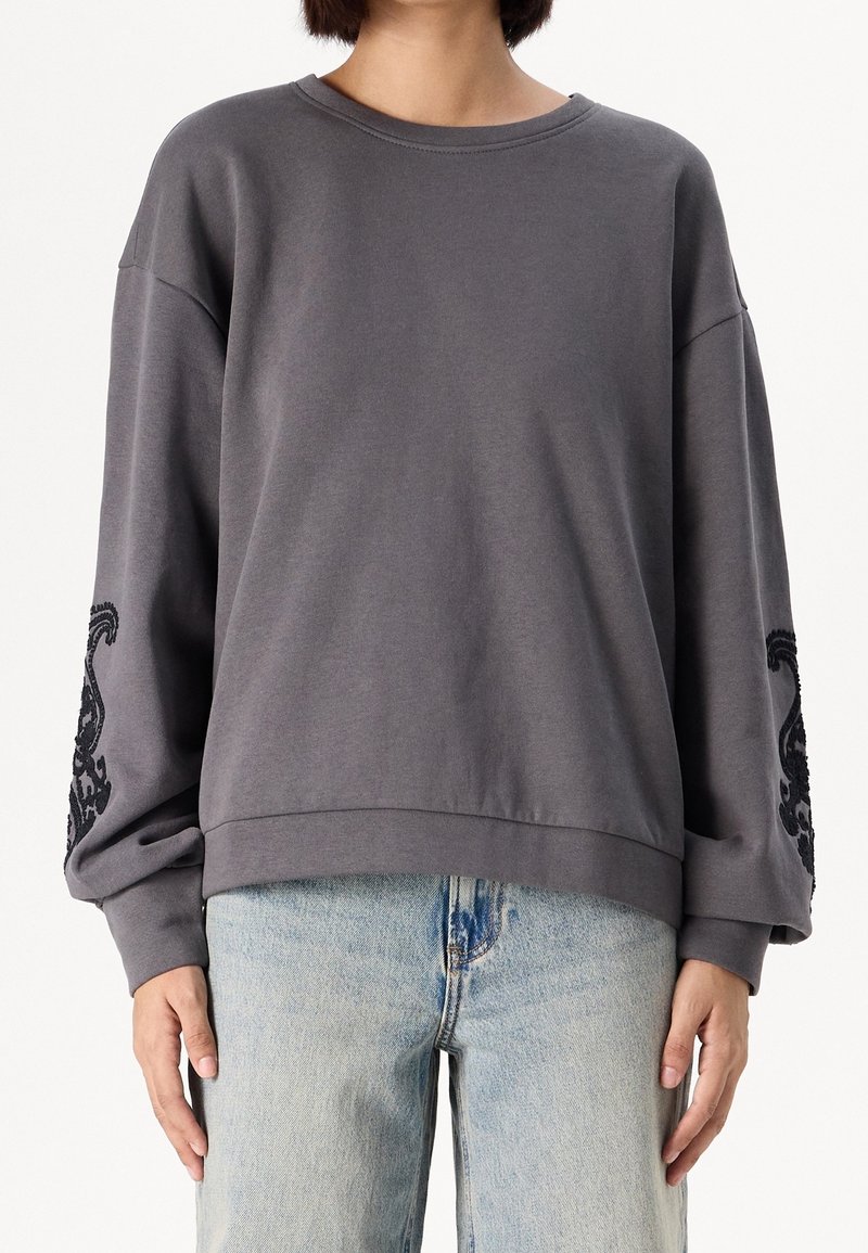 Personne portant un sweat-shirt ample gris foncé avec une broderie noire sur les manches et un jean bleu clair, debout, les bras détendus le long du corps.