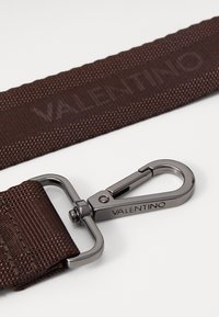 Rjav tekstilni trak z teksturirano površino; vključuje kovinski sponko in odtisnjen znak "VALENTINO".