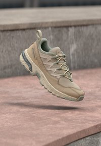 Beige und grüne Salomon-Wanderschuhe mit Schnellschnürung in der Luft über braunen Steinstufen, der robuste Sohlen und der Zugschlaufe sind zu sehen.