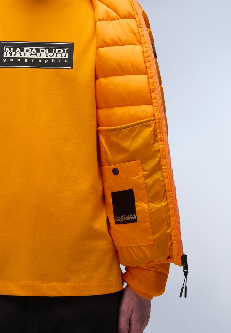 Pomarańczowa kurtka puchowa o gładkiej, błyszczącej fakturze; z ribbowanymi mankietami i zamkniętą kieszenią na ramieniu; nałożona na żółtą koszulkę z logo.
