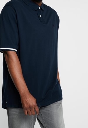 Mężczyzna w granatowej koszulce polo z białym obszyciem na rękawach i jasnoszarych dżinsach, stojący z jedną ręką w kieszeni.