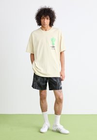 T-shirt oversize beige chiaro con stampa di un cactus verde, abbinata a pantaloni corti neri a fantasia e scarpe da ginnastica e calze bianche.