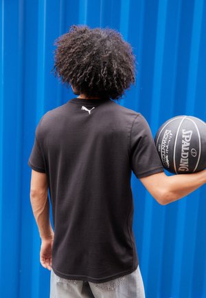 Osoba z kręconymi włosami trzymająca czarną piłkę do koszykówki Spalding, ubrana w czarną koszulkę Puma, stojąca na tle niebieskiej, falistej metalowej ściany.