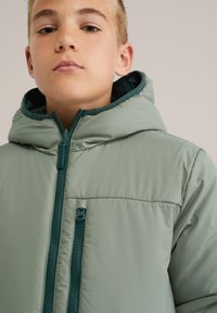 Zelena pufa jakna s kapuco, ki ima sprednje zadrgo in kontrastno temno zeleni notranjosti. Gladka textura in lahka zasnova.