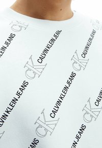 Felpa bianca in cotone con colletto a girocollo, caratterizzata da una ripetizione di testo e logo "Calvin Klein Jeans" neri in diverse dimensioni su tutta la superficie.