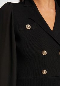 Blouse noire à côtes avec décolleté en V, design à double boutonnage et boutons dorés avec des détails élaborés. Texture douce avec des épaules structurées.