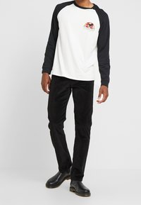 Witte lange mouwen shirt met zwarte raglanmouwen, voorzien van een kleurrijke graphic en logo. Gecombineerd met zwarte ribfluwelen broek en zwarte laarzen.