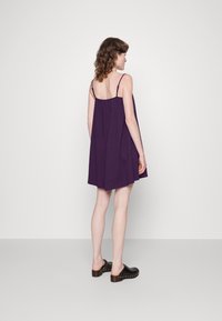 Vestido roxo sem mangas com um corte solto, a cair até metade da coxa, com alças finas e um costurado nas costas. Combinado com tamancos pretos.