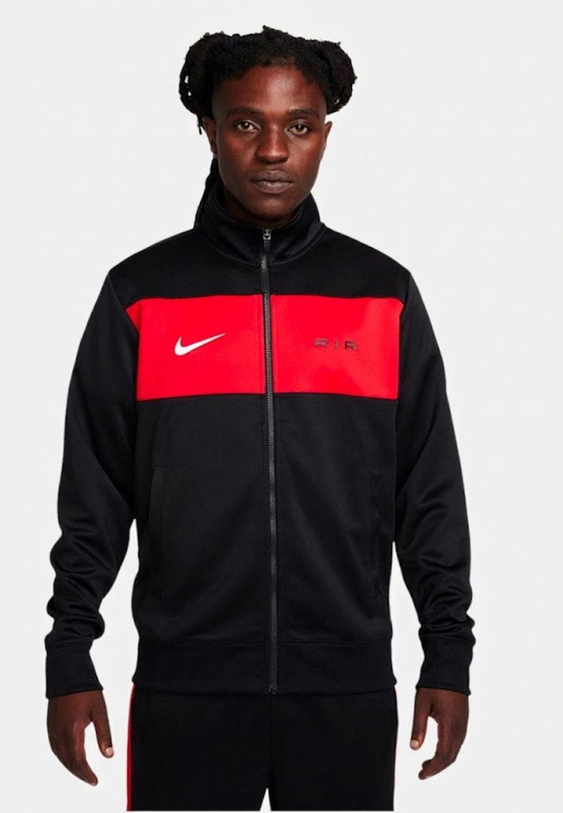 Mann trägt eine schwarz-rote Nike-Zipjacke mit hohem Kragen, steht vor einem schlichten weißen Hintergrund.