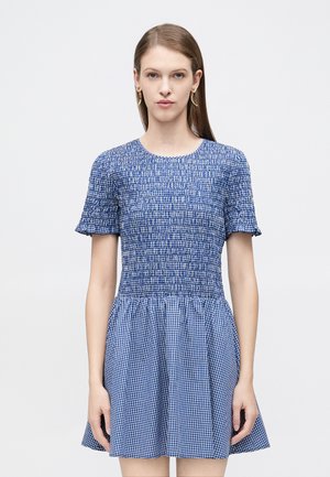 Kvinde iført en blå- og hvidternet kortærmet kjole med rynket overdel og udvidet nederdel, stående mod en ensfarvet hvid baggrund.