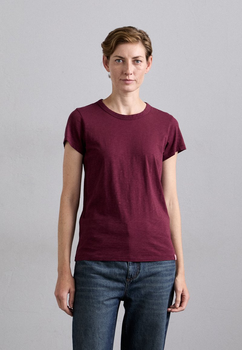 Burgunderfarbene Baumwoll-T-Shirt mit Rundhalsausschnitt und kurzen, umgeschlagenen Ärmeln, kombiniert mit blauen Jeans. Minimalistisches Design, glatte Textur.