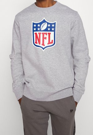 Person in einem hellgrauen Sweatshirt mit rotem, weißem und blauem NFL-Logo und dunklen grauen Hosen, mit den Händen in den Taschen.