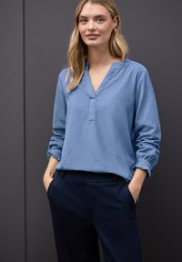 Camicia azzurra a maniche lunghe con scollo a V e polsini arricciati, abbinata a pantaloni scuri; texture del tessuto liscia, design informale.