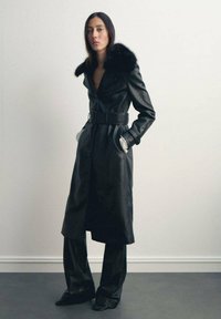 Cappotto lungo in pelle nera con colletto in pelliccia, vita cintata e maniche lunghe. Abbinato a pantaloni neri coordinati e scarpe a punta.