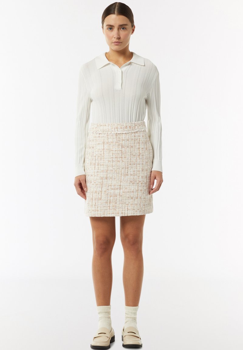 Biała prążkowana koszula z długim rękawem i kołnierzykiem, w połączeniu z beżową mini spódnicą o fakturze i kolorowymi włóknami. Modelka ma na sobie beżowe buty i skarpetki.