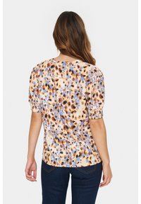 Blouse à imprimé floral avec des manches courtes bouffantes, un col rond et un tissu texturé dans des nuances de marron et de bleu sur un fond beige.