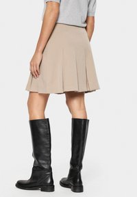 Beige pleated skirt valmistettu pehmeästä kankaasta, polvipituinen, yhdistettynä mustiin polven ylle ulottuviin nahkasaappaisiin, sileä rakenne ja istuva siluetti.