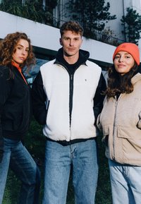 Tre personer poserar utomhus. Nyckelfunktioner: en vit fleece zip-up väst, en svart zip-up jacka och en beige fleece ytterkläder; avslappnade blå jeans.